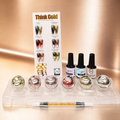 A nail polish set displayed on a clear acrylic stand against a warm metallic gold background. The set includes four bottles of nail polish with black caps, five round clear jars showing different metallic shades, a white instruction card titled 'Think Gold,' and a dual-ended applicator wand with gold glitter polish visible inside.