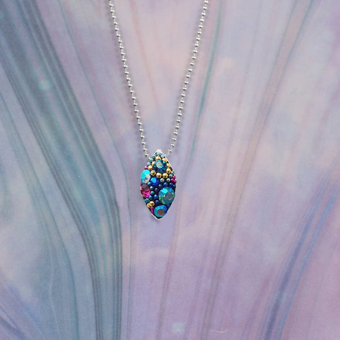 Bijou Seed Necklace by Mosaico Jewels showcased with colorful crystals on a seed-shaped pendant, surrounded by soft pastel hues.