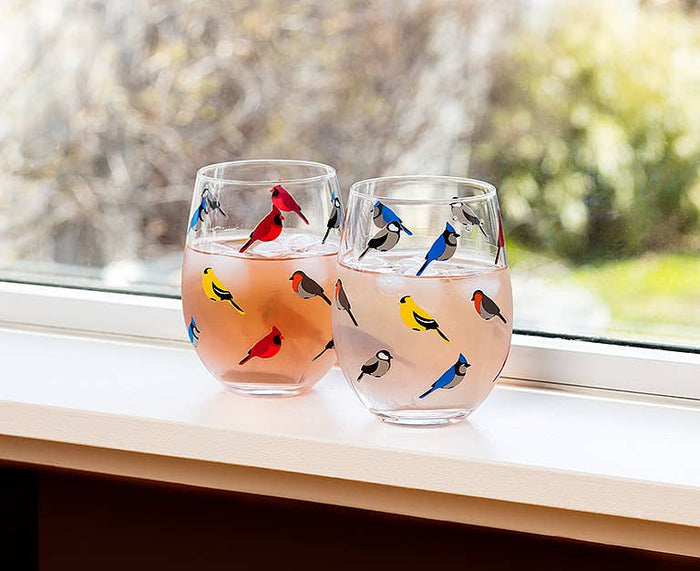 Multi Bird Stemless Wine Glass by Abbott placed on a window sill, displaying a charming assortment of colorful birds against a natural backdrop.