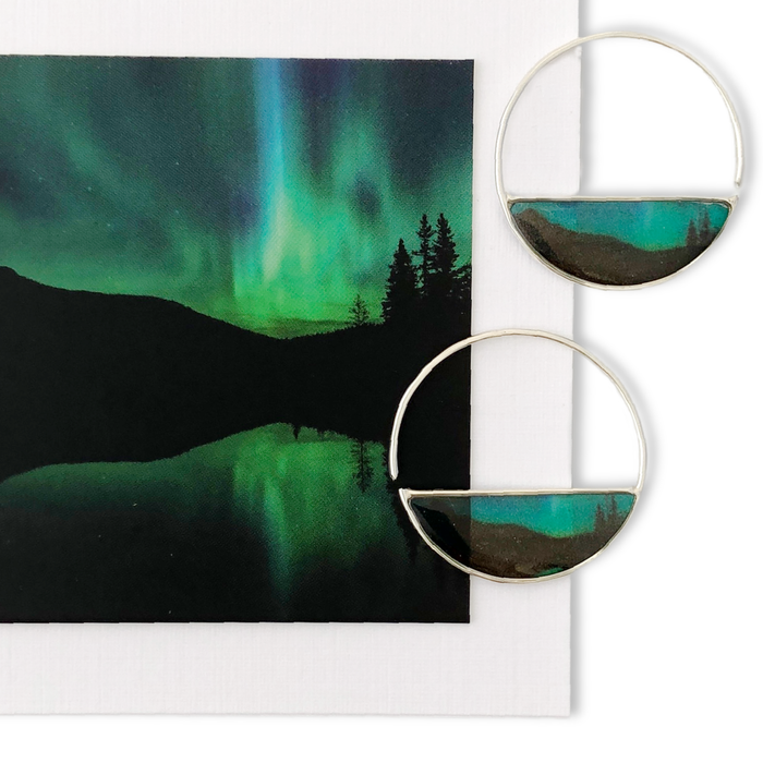 Northern Lights Rise To Kiss The Sky Hoop Earrings by Foterra Jewelry, featuring handmade crescent-shaped design in .925 sterling silver, displayed alongside a picturesque image of the Northern Lights reflecting on water.